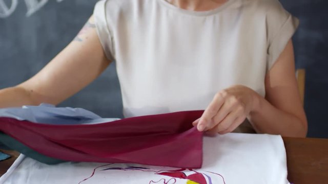 Tilt down shot of creative woman with short blond hair choosing chiffon fabric for decoration of t-shirt with hand-painted image of abstract face