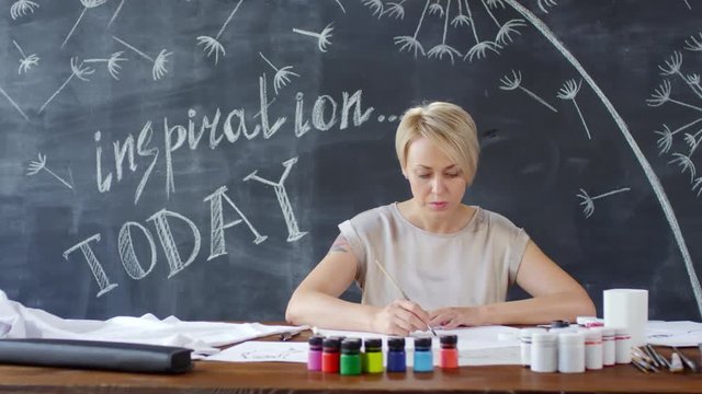 PAN shot of female artist with short blond hair sitting at wooden table in art studio with chalkboard mural and painting on paper with acrylics