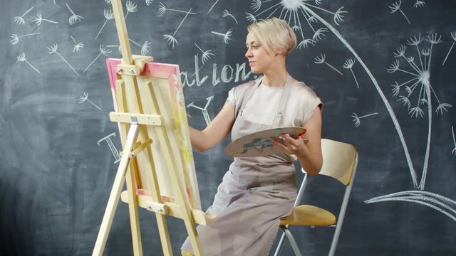 Medium shot of happy female artist wearing apron sitting on chair and painting on easel in art studio