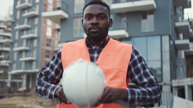 Handsome serious Afro-American builder walks down the construction site and puts on the white hard hat. Safety first, profession concept. Low angle shot, male portrait