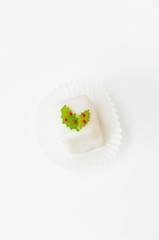 Fototapeta na wymiar White Christmas petit four, small cake square, covered with sugar icing and decorated fondant ilex leaves and red berries in paper wrapper on white background