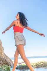 Portrait of latino woman with curly hair, red top and denim shorts running at beach