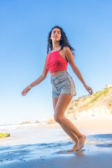 Portrait of latino woman with curly hair, red top and denim shorts running at beach