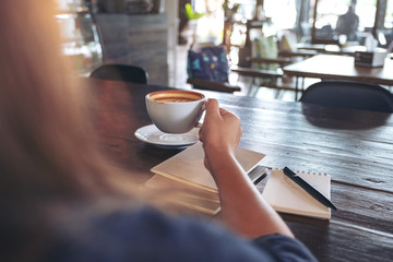Closeup image of a woman holding and drinking hot coffee with notebooks on table in cafe