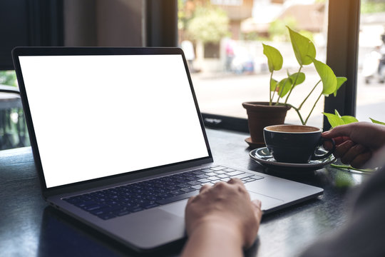 Mockup image of a woman's hands using and touching laptop touchpad with blank white desktop screen while drinking coffee on wooden table in cafe