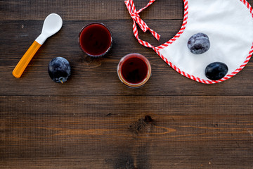 Fruit puree for small babies. Puree with prunes in bowl near punes, spoon, bib on dark wooden background top view space for text