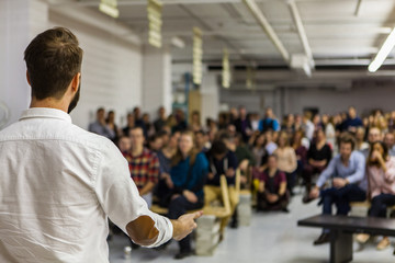 Man with white shirt is giving a conference in front of 200 people in an industrial environment -...
