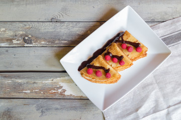 Crepes with fresh raspberries and chocolate syrop on aged wooden background. Top view, copy space.