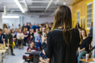 Young woman is giving a conference in front of 200 people in an industrial environment - back view - Blurred audience mainly composed of young adults