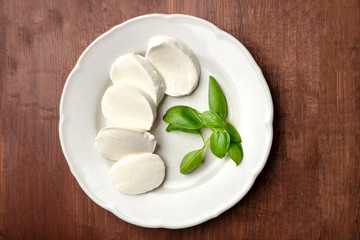 A photo of a white plate with fresh basil leaves and Mozzarella cheese, shot from the top on a dark rustic wooden background with copy space