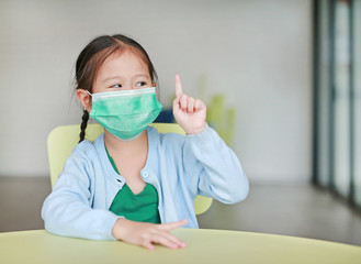 Cute little Asian child girl wearing a protective mask with showing one forefinger sitting on kid chair in children room.