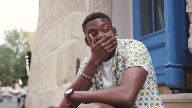 Sad and tired young African man sitting near building with blue door, touching head. Somebody giving smartphone to guy. Happiness. Important message.