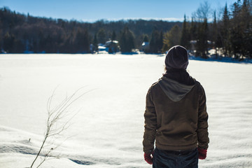 Young man wearing a tuque is contemplating the horizon on the side of a fronzen lake in the winter time - Back view with blurry valley in the background