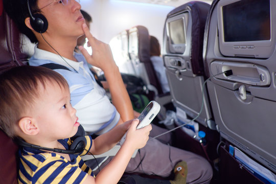 Curious Asian 2 years old toddler use remote controls to change display options of computer on seat during flight on airplane with father, Entertainment in flying aircraft, Little traveler concept