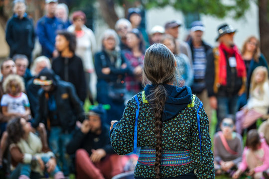 Woman with platted hair and glasses is giving a public speech or conference at the park in front of a blurry crowd of 50 people