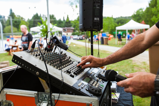 DJ Is Adjusting The Levels On His Mixing Board During A Community Event At The Park - Lateral View With Blurry People Talking And A Marquee In The Background