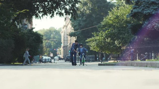 Nice child riding a bike through the street, father helping and walking near guy in park. Good-looking Caucasian man with beard looking kid riding away