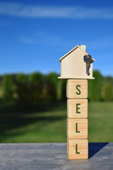 Sell - word on wooden cubes and house model with key on green blurred landscape and blue sky background with copy space for text. Real estate sale and moving in new home concept.