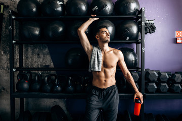 Fototapeta na wymiar Athletic man with a naked torso and t-shirt on his shoulder dressed in the black shorts stands near the sport equipment in the gym