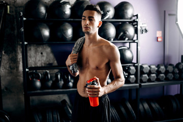 Fototapeta na wymiar Athletic man with a naked torso keeps t-shirt on his shoulder and holds bottle with water in the hand near the sport equipment in the gym
