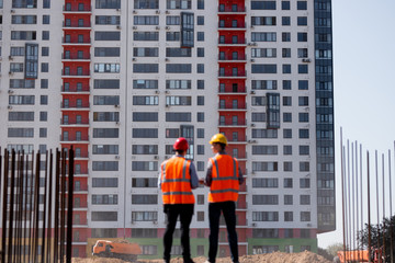 Two civil engineers  dressed in orange work vests and helmets talk about  the construction process on the building site against the background of a multi-storey building