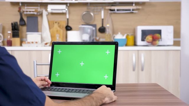 Man at the kitchen looking at laptop with green screen mock up. The chroma has mark for easy keying. Dolly slider 4K footage