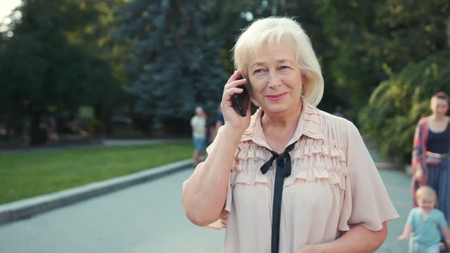 Cheerful old Caucasian lady holding smartphone in hands, touching screen. Lovely grandmother calling daughter. Warm weather. Summertime. Park.