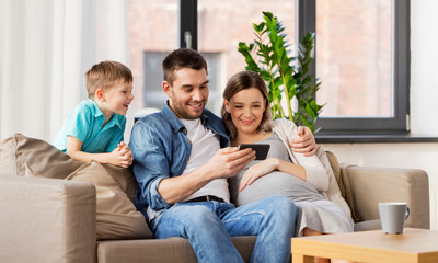 Obraz premium family, pregnancy and technology concept - happy pregnant mother, father and little son with smartphone sitting on on sofa at home
