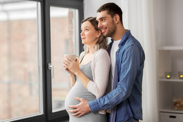 pregnancy and people concept - happy man hugging his pregnant wife standing at window at home