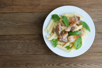 Cooked penne pasta isolated in white round plate, with basil leaves and pork meat, over wooden table, decorated with basil leaves. Top view.
