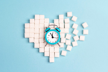 Broken heart made of sugar cubes with alarm clock on a blue background.