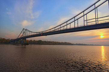 Fototapeta premium Wide angle landscape panorama of Dnipro River and Pedestrian Bridge to the Trukhanov Island. Colorful vibrant sky, sun reflected in the water. Foggy autumn morning during sunrise. Kyiv, Ukraine