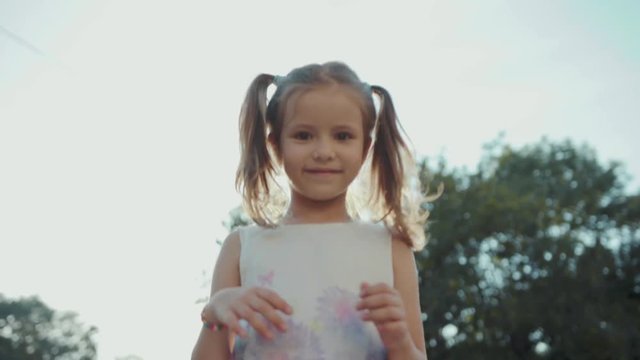 Nice Caucasian litte girl running through the park. Cute Slavic girl with brown eyes stopping, looking at camera, smiling. Hairstyle. Summer's park with people