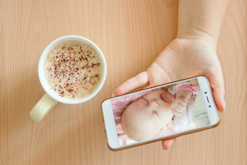 view handheld color video baby monitor. Female hands are holding a smartphone with a baby monitor app. Near hot drink