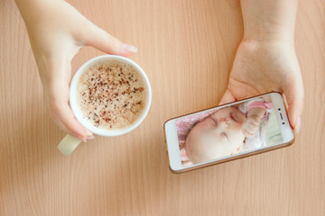 view handheld color video baby monitor. Female hands are holding a smartphone with a baby monitor app. Near hot drink