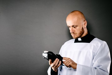 Priest in white surplice and black shirt with white collar holding credit card terminal.