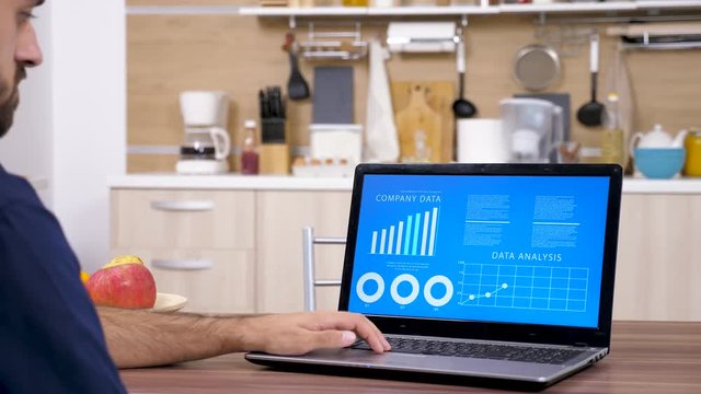 Man looking at animated financial charts and data at the table on his laptop. There is a cozy and modern kitchen in background. Dolly slider 4K footage
