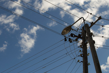 Electric wires hanging on a pole with a lighting lamp photographed from below on a background of blue sky with white clouds
