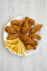 Tasty fried chicken drumsticks, spicy wings, French fries and chicken fingers on white plate over white wooden background, top view. Flat lay, overhead, from above.