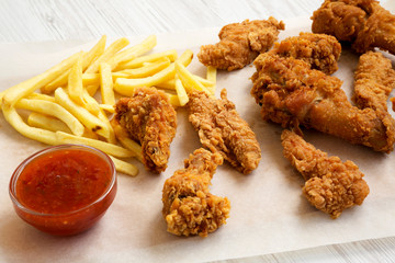 Delicious fast food: fried chicken legs, spicy wings, French fries and chicken fingers with sour-sweet sauce on baking sheet over white wooden background, close-up.