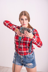 Young sexy woman stands near the white wall and holds a drill. Girl in shorts and a plaid shirt...