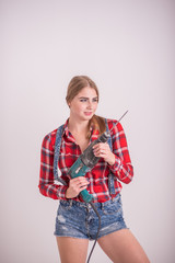 Young sexy woman stands near the white wall and holds a drill. The girl in shorts and a plaid shirt...