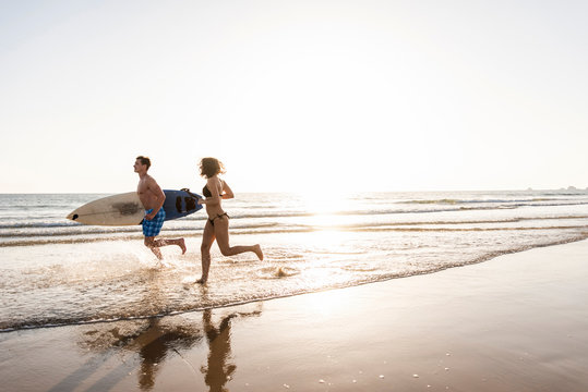 Young couple running on beach, carrying surfboard
