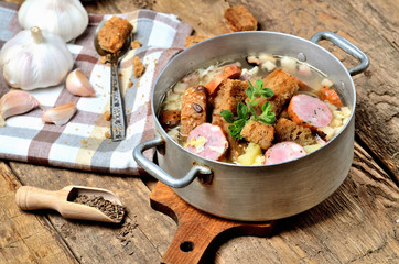 Old saucepan with garlic soup, fresh garlic bulbs, cloves, colored pepper, wooden spoon, croutons, lable and a towel on a rustic wooden table