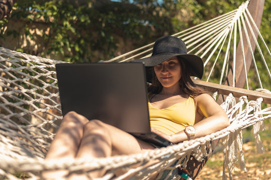 Brunette girl in a yellow swimsuit sitting in a hammock using a laptop, work on vacation, freelance, remote work, online earnings