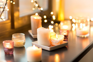 hygge, decoration and christmas concept - candles burning in lanterns and festive garland on window...