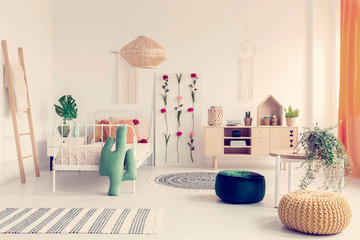 Boho kid's bedroom with white metal bed, wooden furniture and colorful poufs, real photo