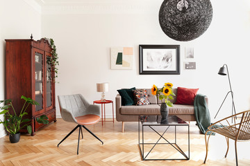Sunflowers on table between armchairs in white flat interior with posters above sofa. Real photo