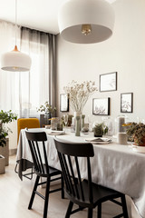 Lamps above black chairs at table with flowers in bright dining room interior with posters. Real photo