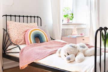 Plush toy, rainbow pillow and pink blanket on kid's bed in white bedroom interior. Real photo - obrazy, fototapety, plakaty